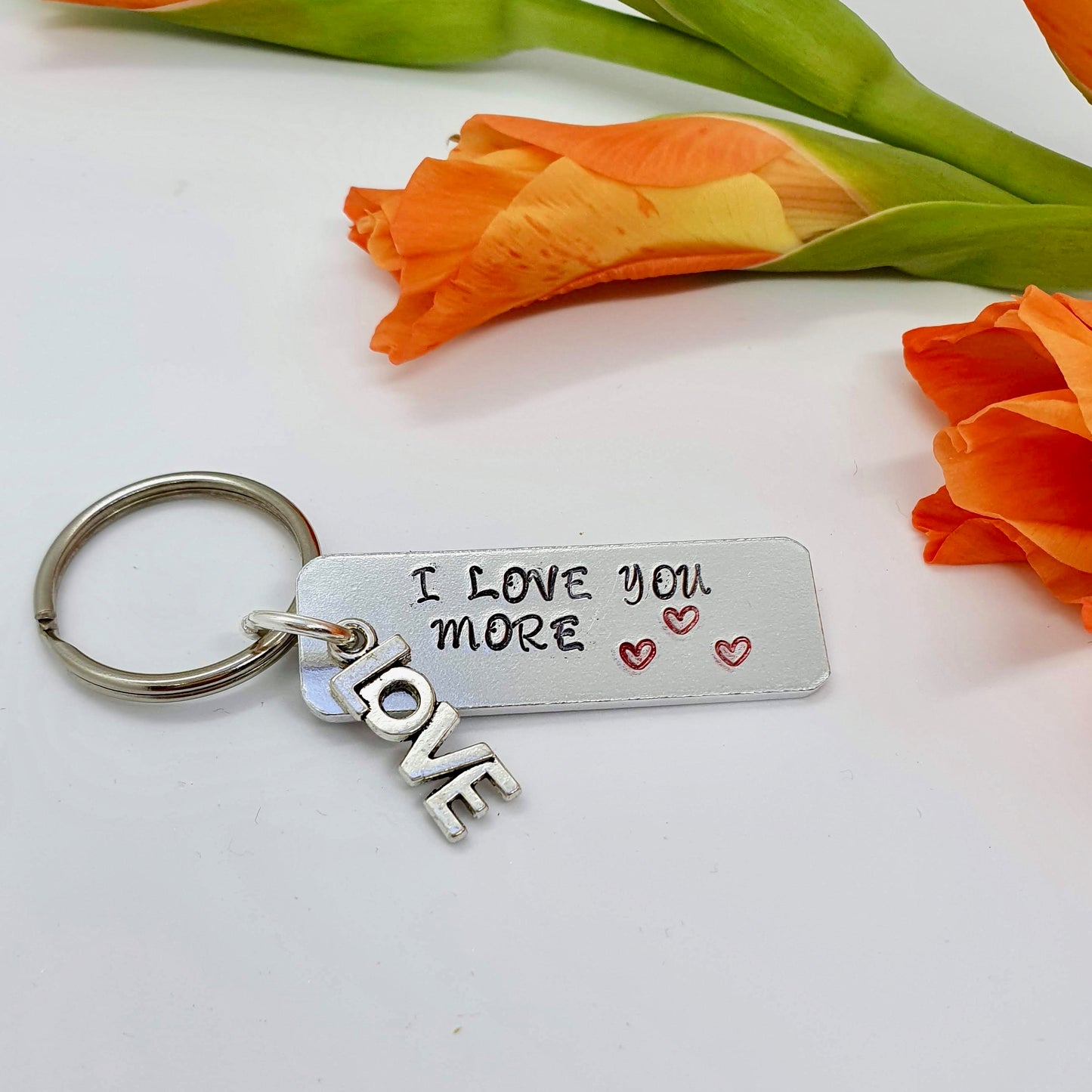 Aluminium rectangular  keyring with 'I love you more' and 3 red hearts hand stamped on it. A separate charm with the word 'Love' is also attached. 